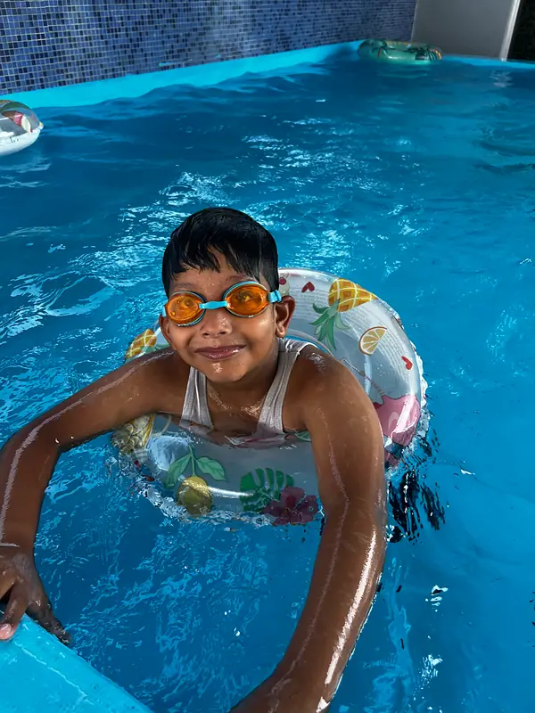 A boy in orange goggles grinning from his swim ring at the summer camp pool