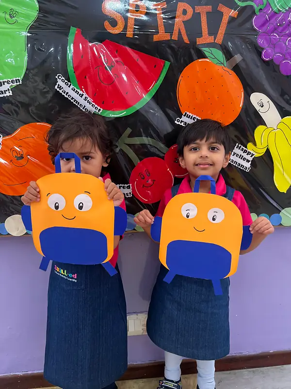 Two friends holding their backpack crafts in front of the fruit wall
