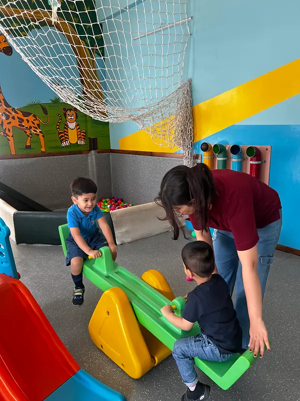 A teacher helps two children on the seesaw in the indoor play area