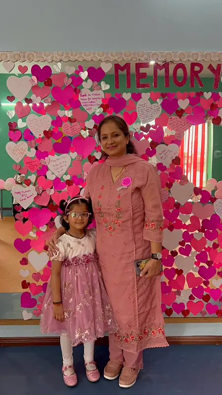 A mother and daughter smile in front of the Mother's Day memory wall of hearts